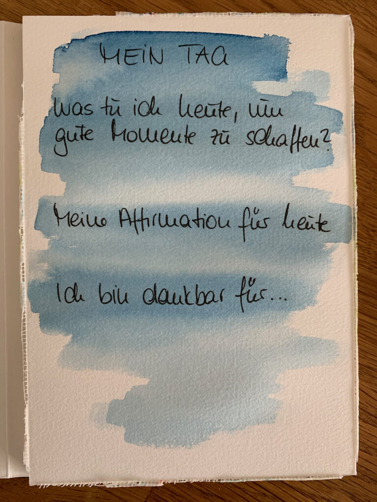 Aquarellpapier mit Text, hinterlegt mit blauer Aquarellfarbe. Hier steht: Mein Tag, Was tu ich heute, um gute Momente zu schaffen?, Meine Affirmation für heute, Ich bin dankbar für...