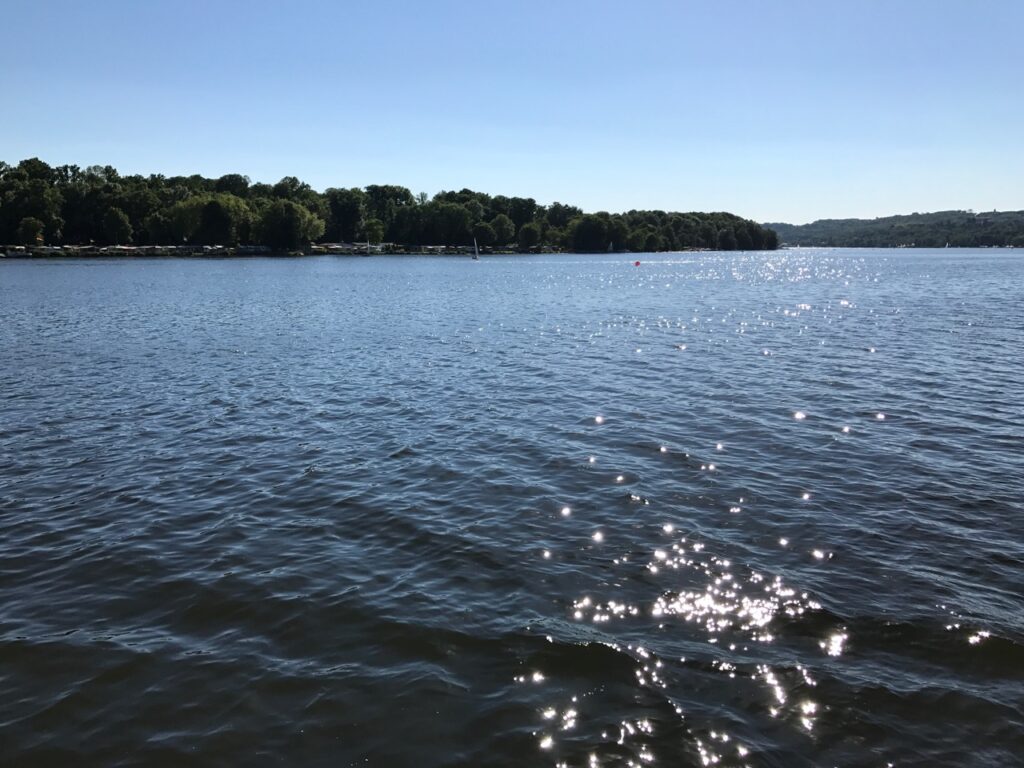 Juli: Glitzerndes Wasser auf einem See. Bäume am Ufer im Hintergrund.
