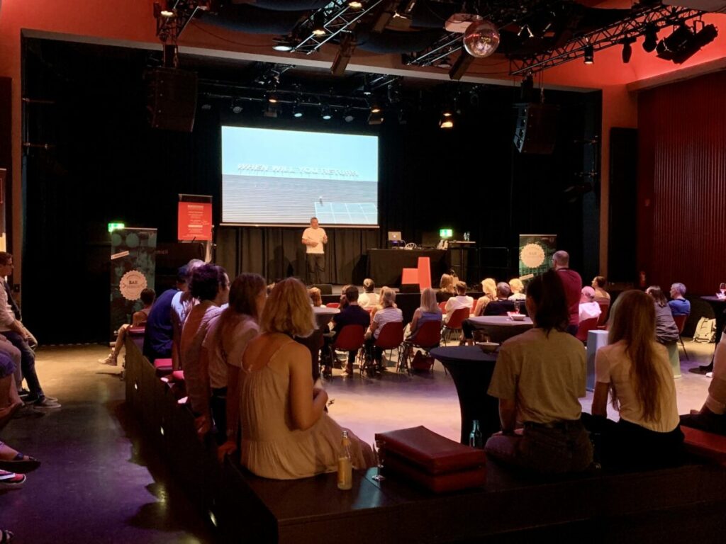 BARsession mit Raphael Gielgen: Raphael Gielgen von Vitra beim Vortrag auf der Bühne. Davor sitzen Gäste der BARsession auf Stühlen bzw. stehen an Stehtischen.