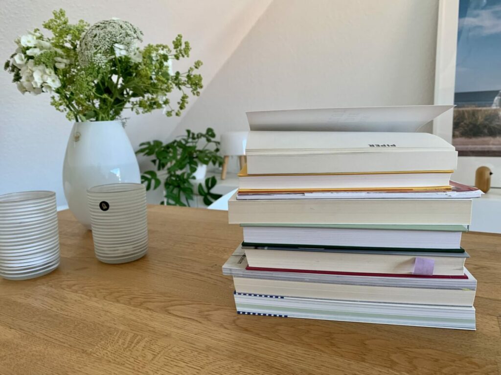 Im Vordergrund Bücherstapel umgedreht, so dass man die Titel nicht lesen kann, dahinter eine weiße Vase mit grün-weißen Blumen