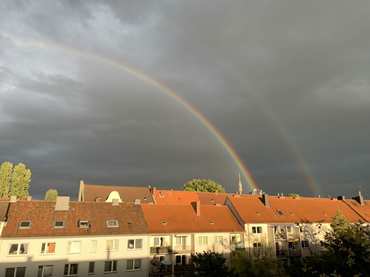 Doppelter Regenbogen: Von der Sonne angestrahlte Häuserreihe vor sehr (!) dunklem Himmel, dahinter zwei Regenbögen