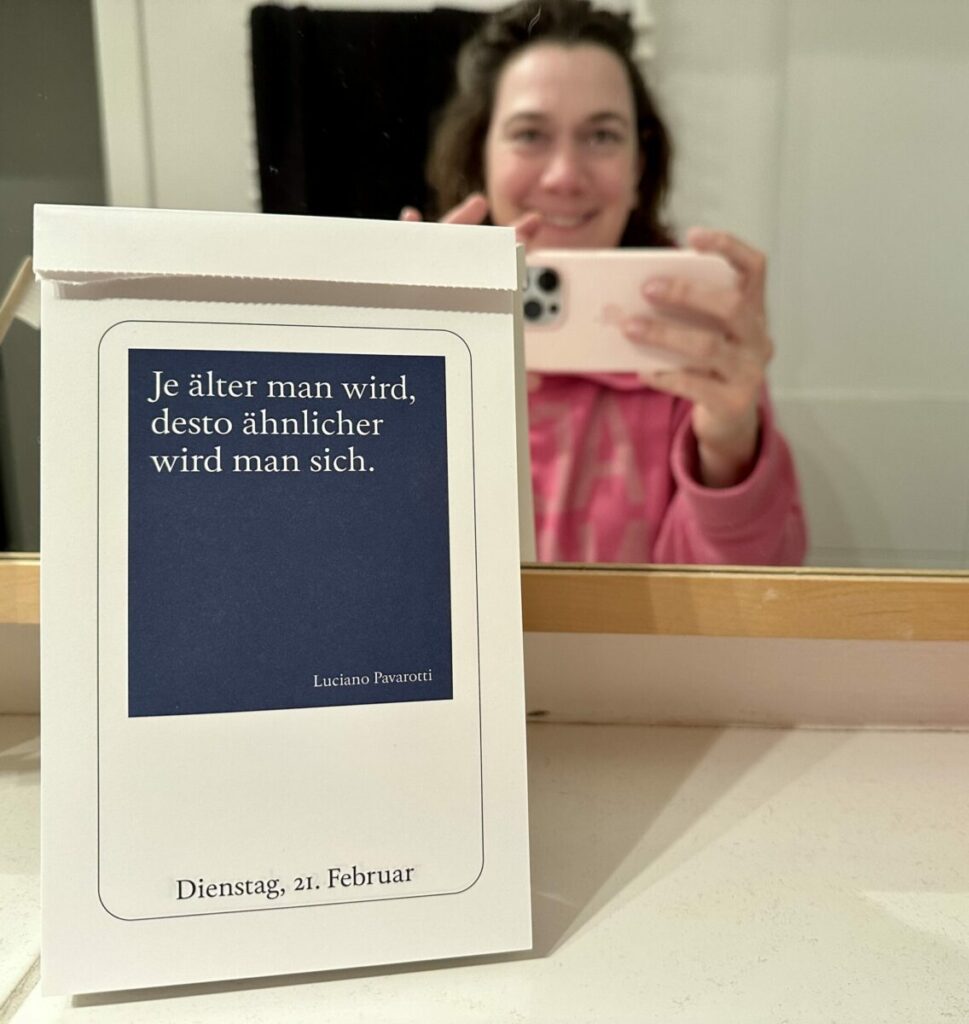 Diogenes-Kalender mit der Seite vom 21. Februar 2023. Zitat: "Je älter man wird, desto. ähnlicher wird man sich." von Luciano Pavarotti. Dahinter im Spiegel die Blogbesitzerin erkennbar.