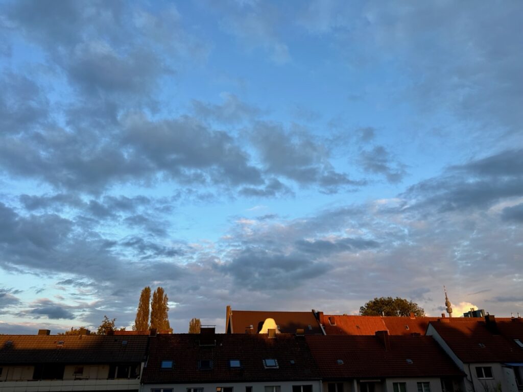Blaue Stunde: Himmel ist noch blau, davor dunkle Wolken und am unteren Bildrand werden Häuserdächer von der Sonne rot angestrahlt 