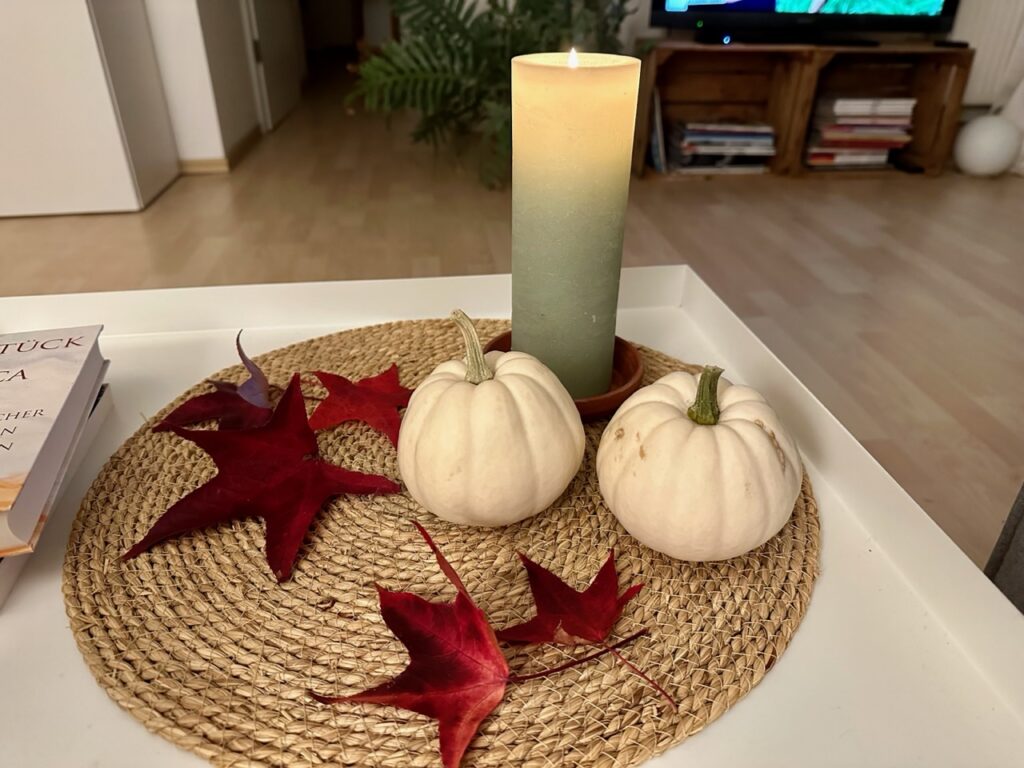 Couchtisch mit runder Strohmatte. Darauf eine brennende Salbei-farbene Kerze mit rotem Laub vom Ahorn und zwei kleinen, weißen Kürbissen