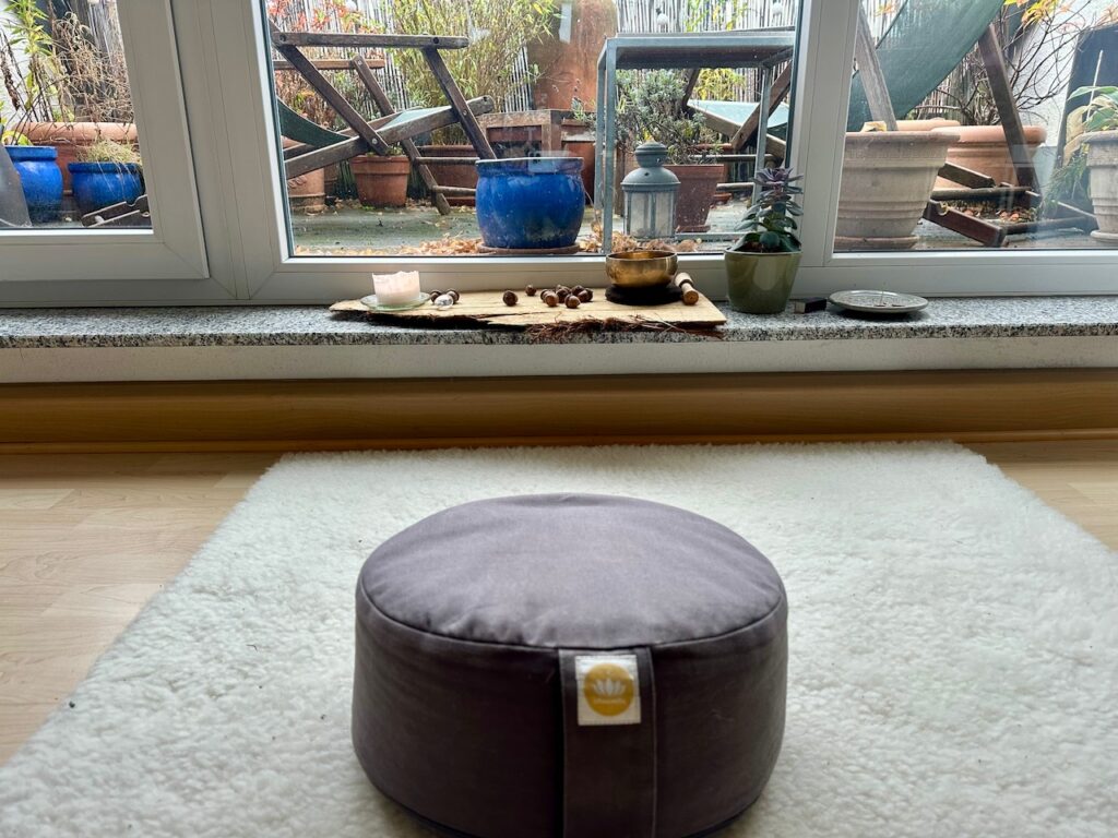 Blick auf ein graues Meditationskissen auf einer Schurwollmatte mit Ausblick auf eine herbstliche Terrasse 