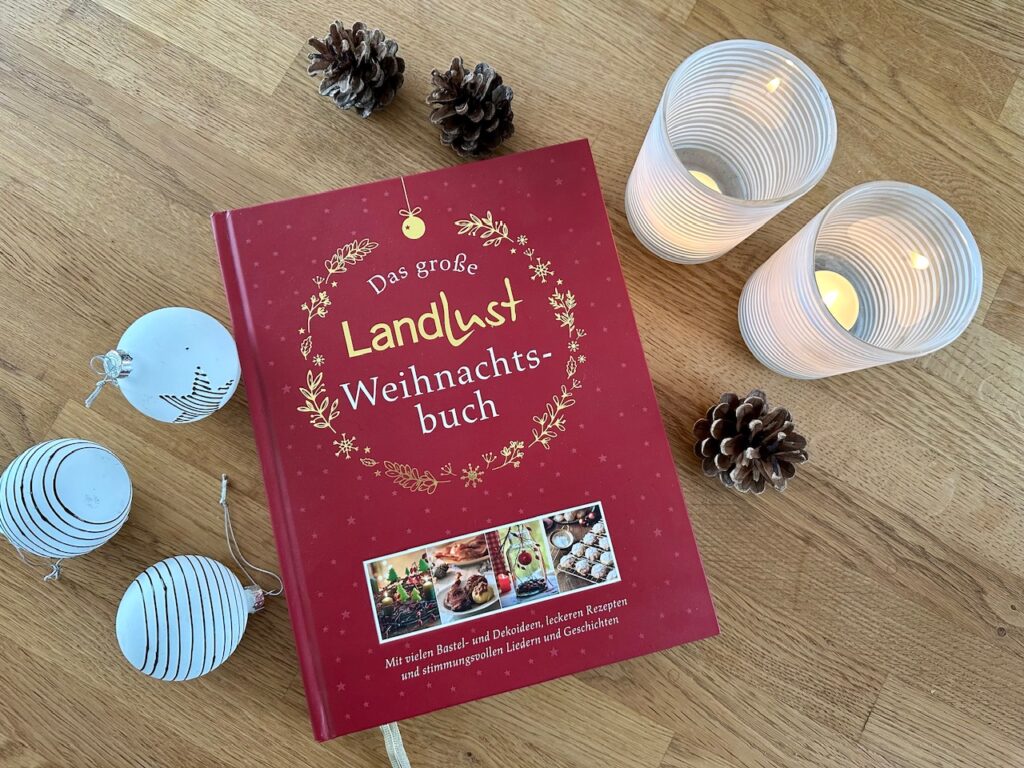 Rotes Buch "Das große Landlust Weihnachtsbuch" liegt auf Holztisch. Darum drapiert zwei weiße Teelichthalter, weiße Christbaumkugeln und Zapfen