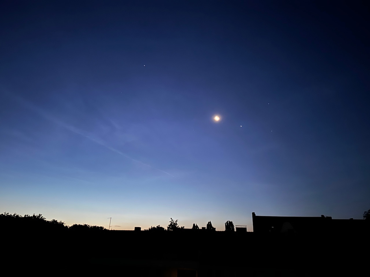 Nachthimmel kurz vor Sonnenaufgang: Unten eine schwarze Silhouette der Häuserdächer, darüber der dunkelblaue Nachthimmel mit einem abnehmenden Mond und diversen Sternen.