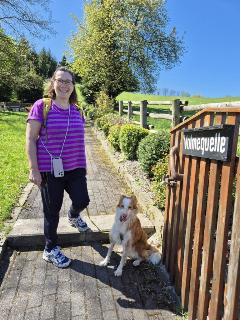 Frau und Hund neben dem kleinen Holztor, wodurch man zur Quelle gelangt. An dem Tor hängt ein Schild "Volmequelle". 