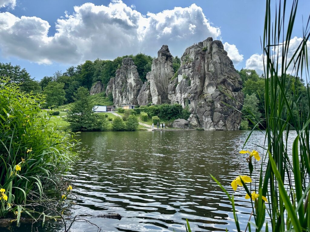 Blick auf die Externsteine bei Blauem Himmel mit Wolken. Im Vorderung ein See, rechts und links Gras und gelbe Blumen bzw. Busch.