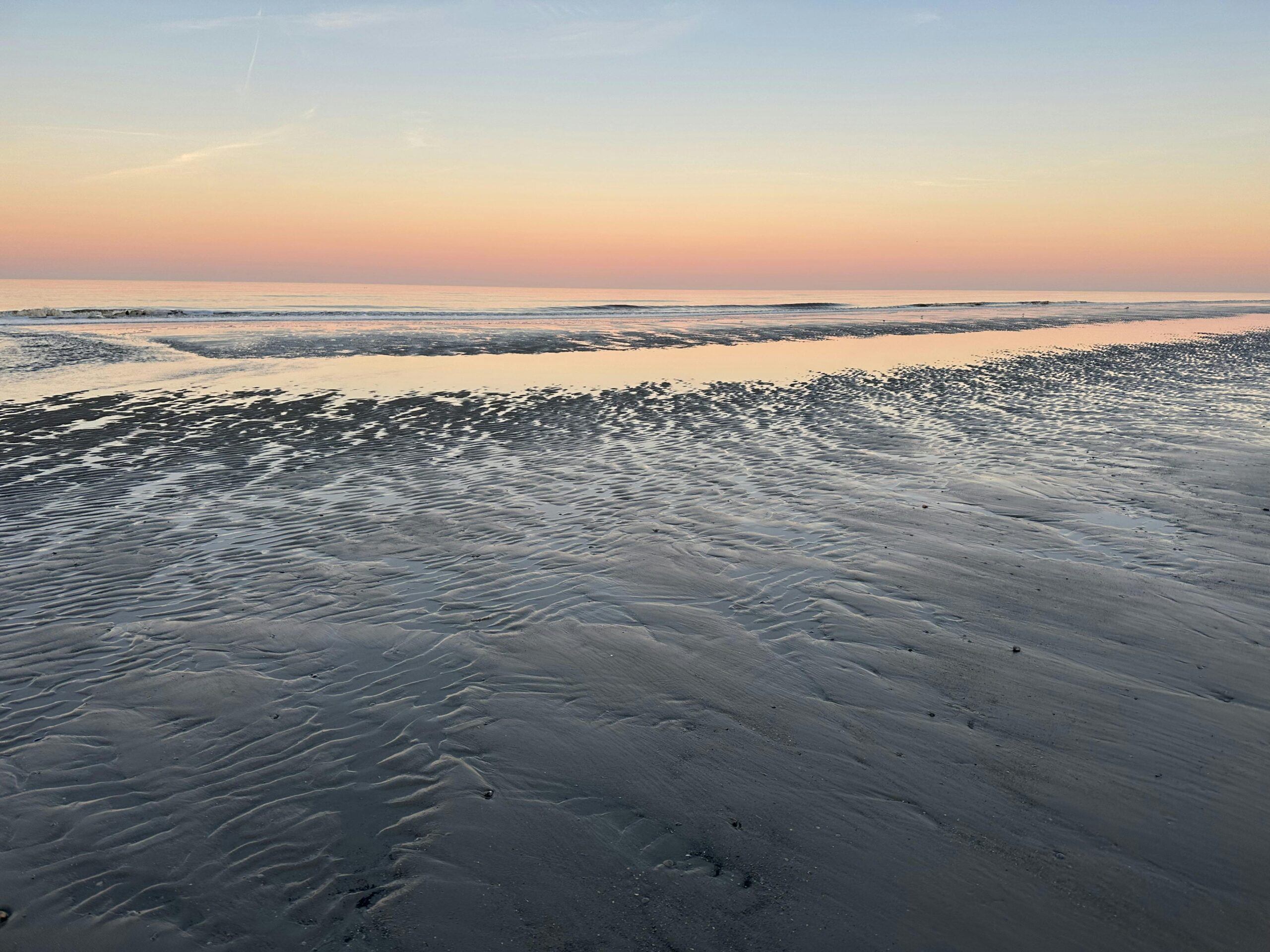 Meersaum bei Sonnenaufgang: Zartrosé- und hellblau-farbener Himmel, der sich im Meer spiegelt.
