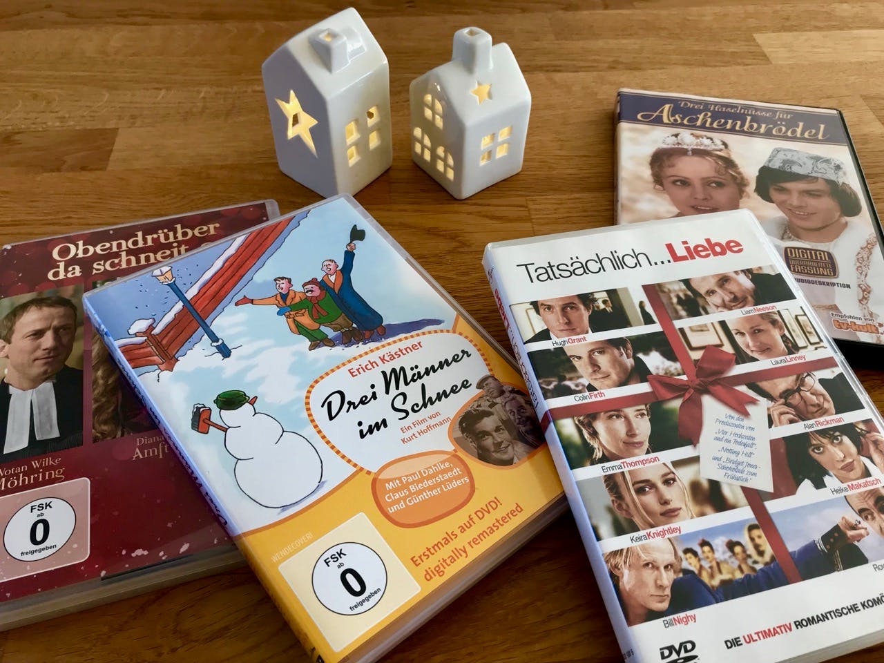 Vier DVD-Hüllen auf einem Holztisch mit zwei kleinen, weißen Porzellan- Häuschen, die leuchten. Die DVDs: "Obendrüber da schneit es", "Drei Männer im Schnee", "Tatsächlich Liebe", "Drei Haselnüsse für Aschenbrödel"