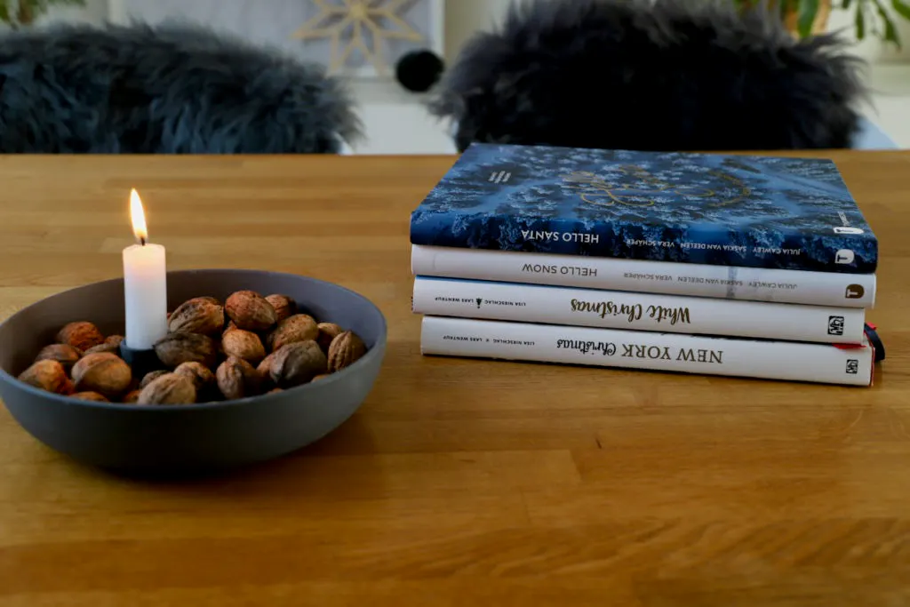 Holztisch. Darauf: Eine Schale mit einer brennenden Kerze darin und Nüssen Daneben ein Stapel Weihnachtsbücher.