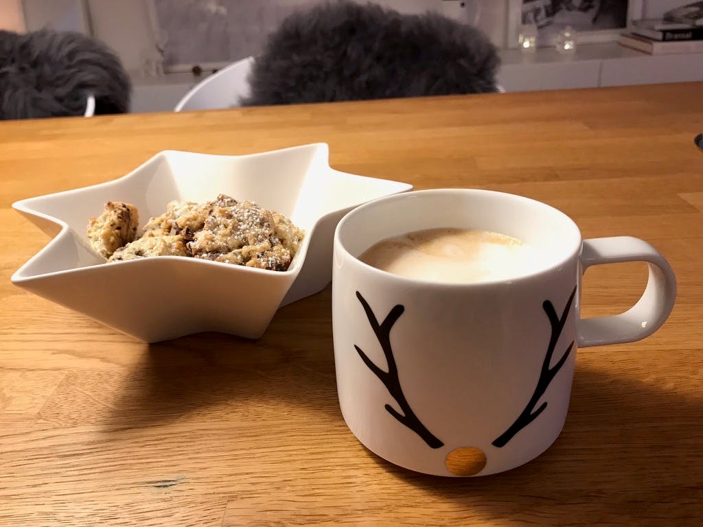 Tasse mit schematisiertem Hirschaufdruck, gefüllt mit Milchkaffee. Dahinter eine Sternförmige Schale mit Keksen darin.