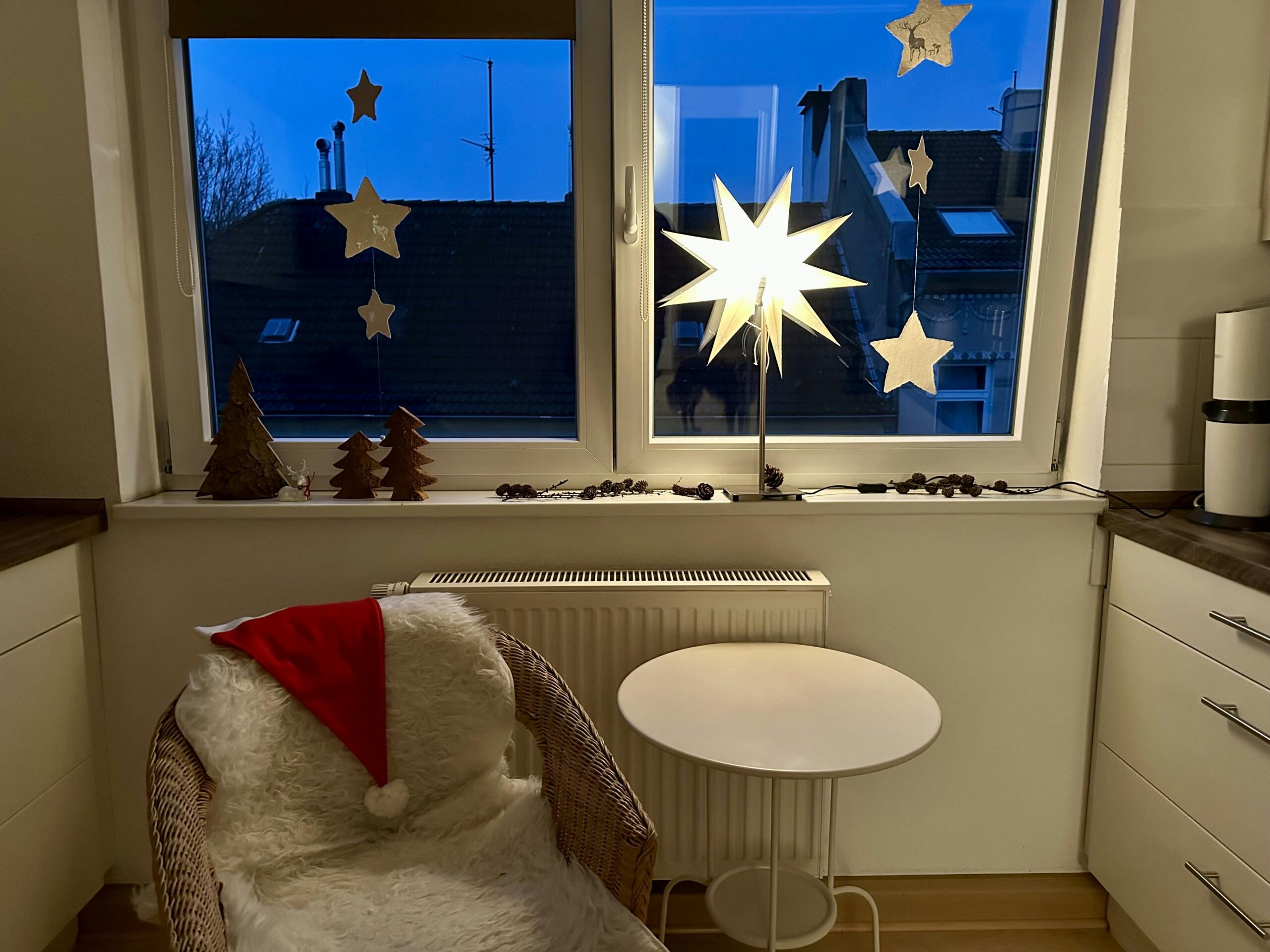 Weihnachtlich dekoriertes Küchenfenster mit Papier-Sternenketten vor dunkel werdendem Himmel. Auf der Fensterbank stehen eine leuchtende Sternlampe, aus Holz gefertigte Tannenbäume sowie kleine Zapfen. Auf dem Korbsessel davor liegen ein helles Fell sowie eine Nikolausmütze.