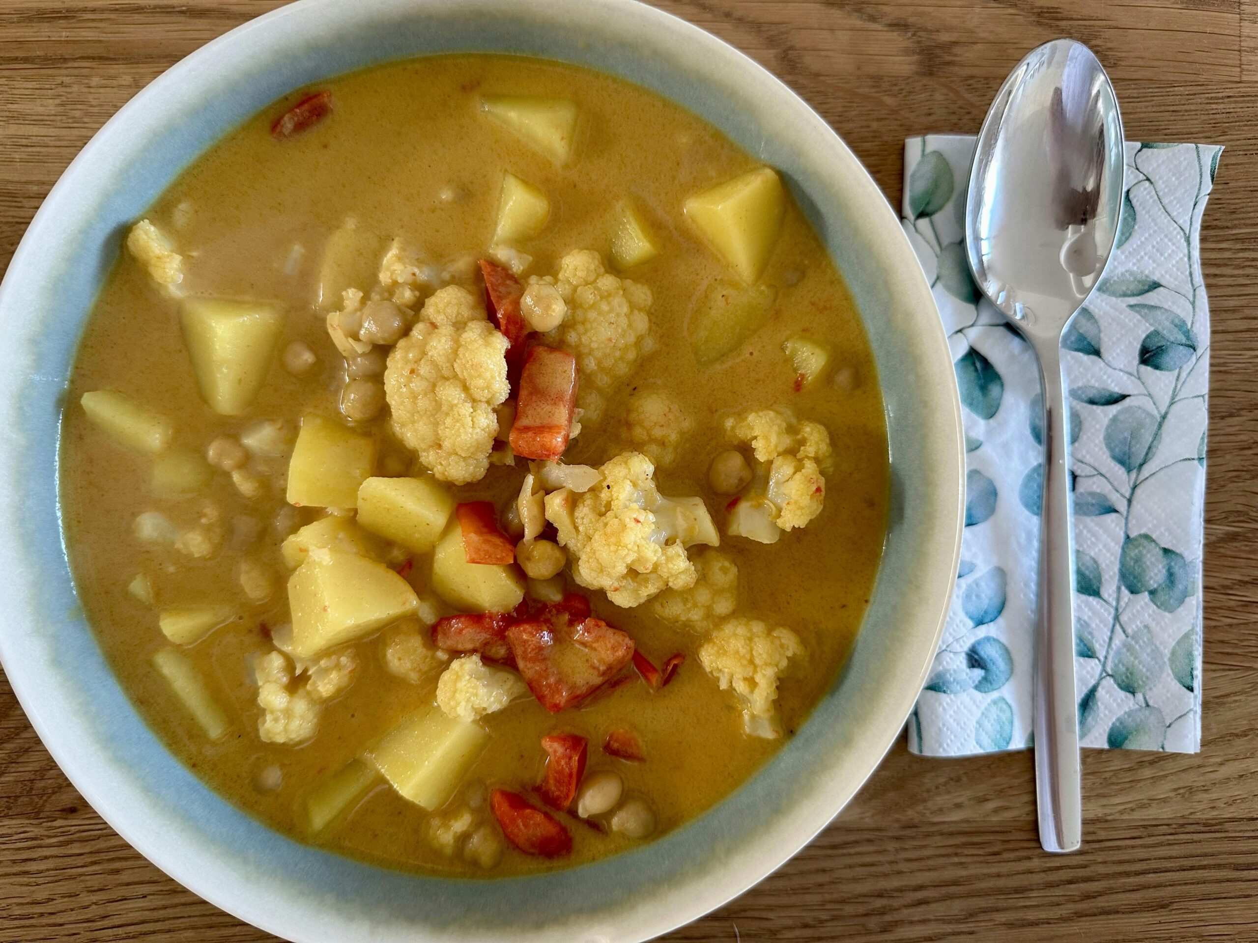 Steingutteller mit einem Curry aus Blumenkohl, Kartoffeln und roter Paprika. Daneben eine Serviette mit einem großen Löffel.