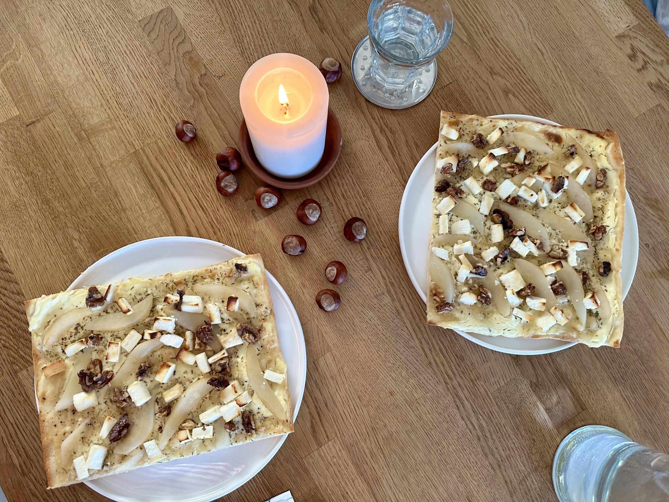 Holztisch mit zwei Tellern mit Flammkuchen, einer brennenden Stumpenkerze und Kastanien.