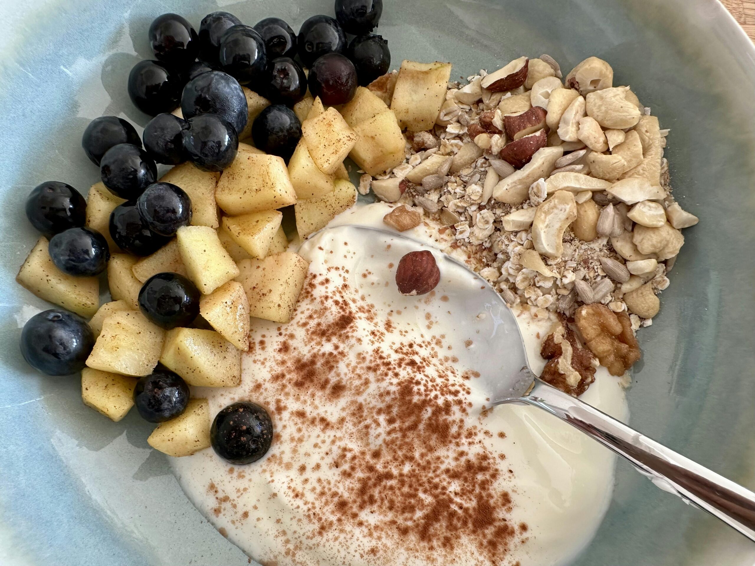 Salbeifarbener Teller mit Naturjoghurt, gebratenen Apfelstücken, Heidelbeeren, Müsli und einem Kakao-Topping.