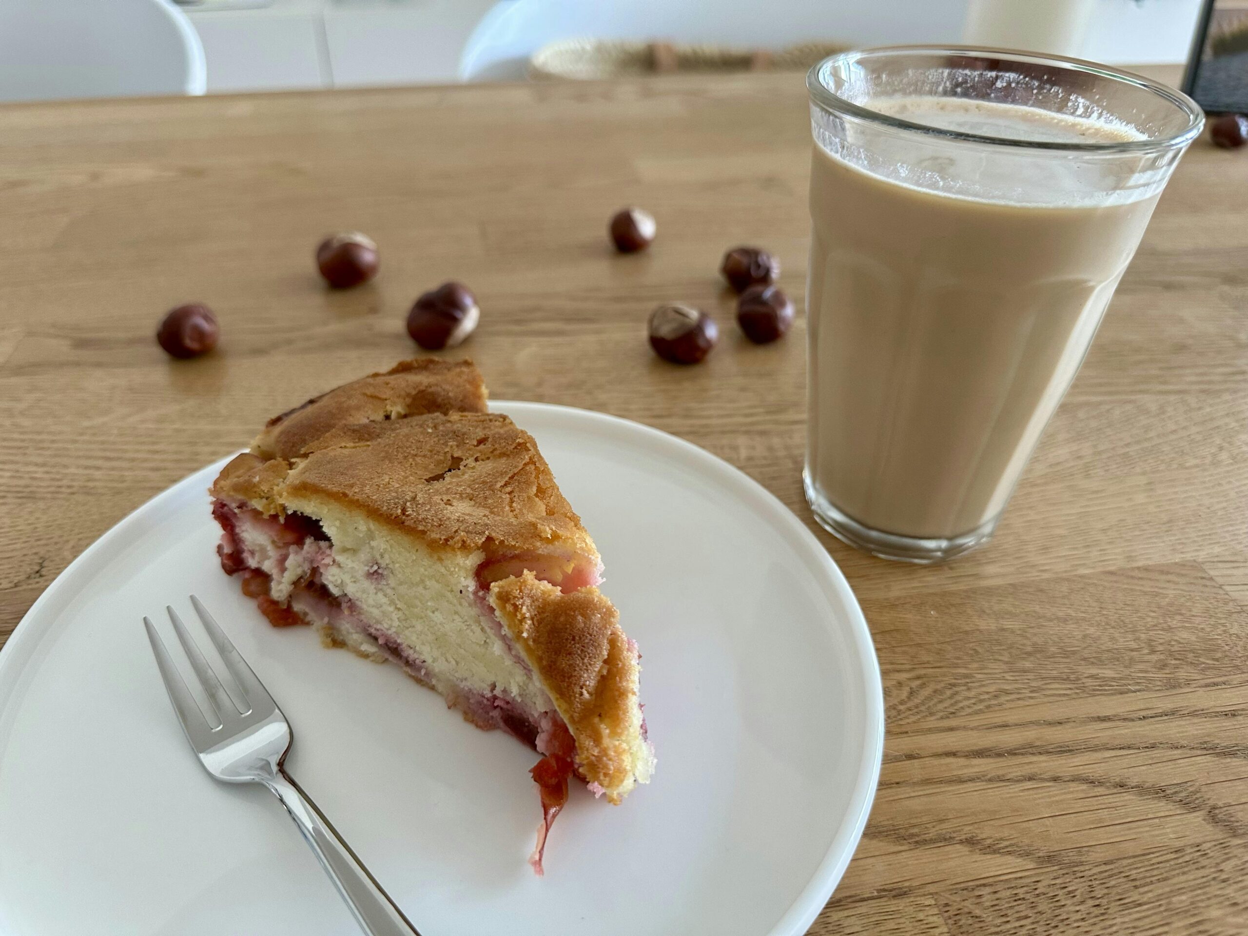 Stück Pflaumenkuchen auf einem weißen Teller und daneben ein Glas mit Latte Macchiato. Im Hintergrund eine Handvoll Kastanien.