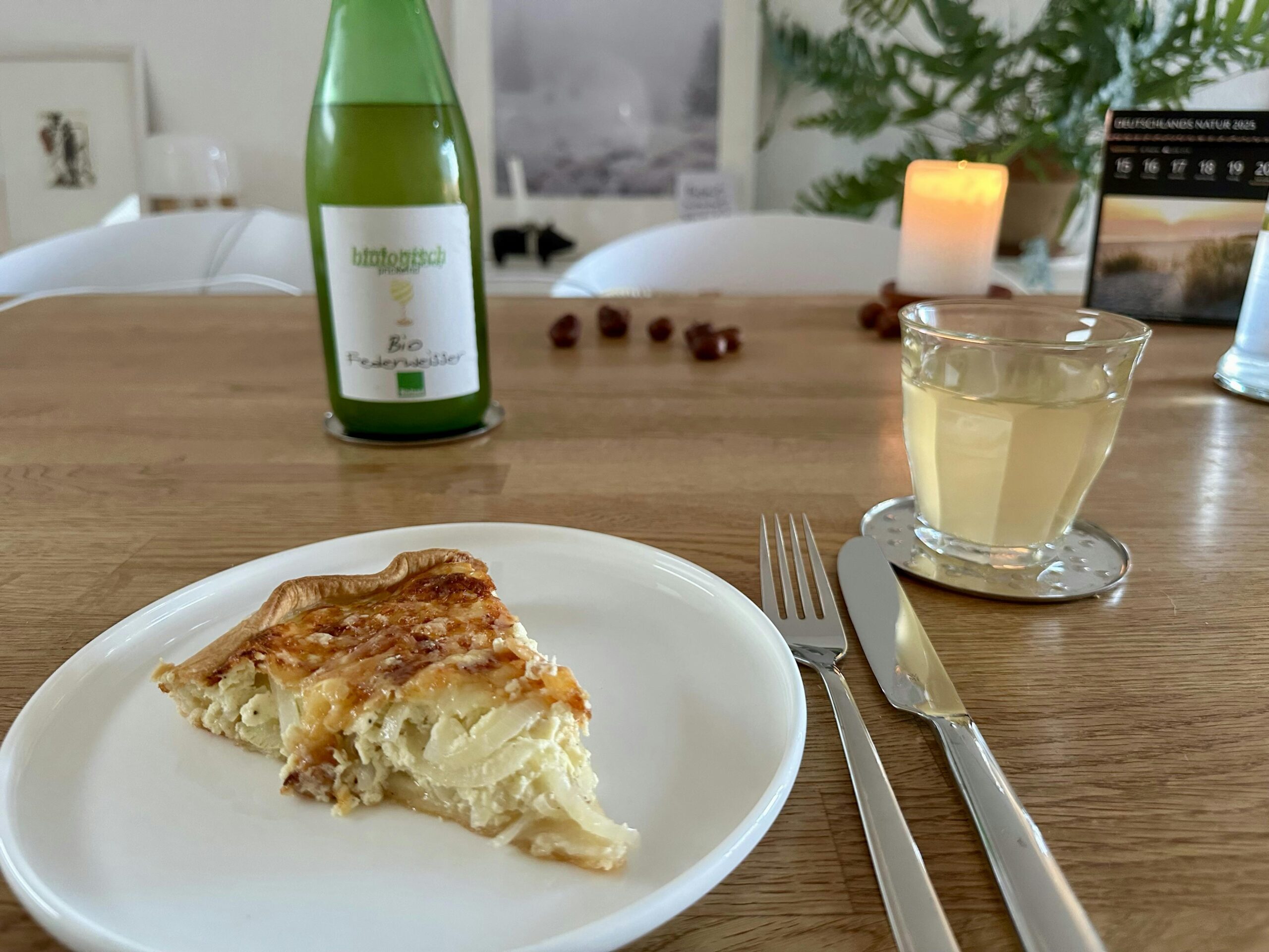 Teller mit einem Stück Zwiebelkuchen, einem Glas Federweißem und im Hintergrund eine Flasche Federweißer und eine brennende Stumpenkerze.