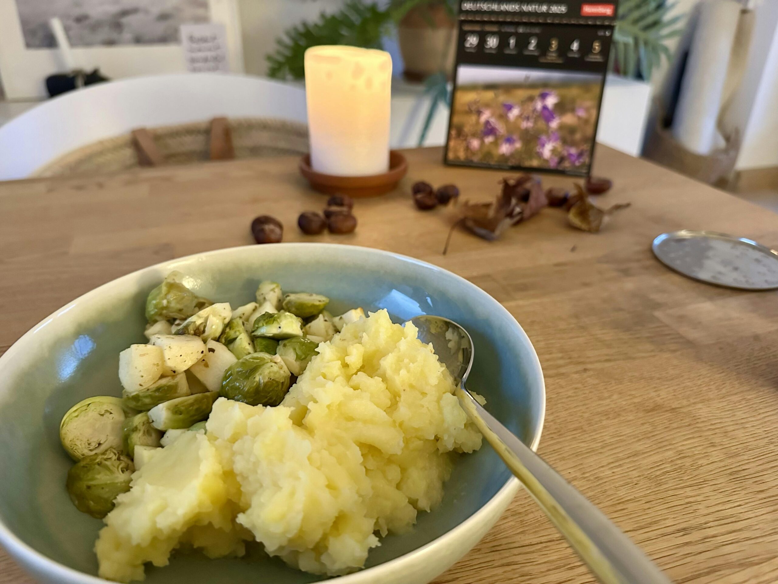 Tiefer, grüner Teller auf einem Esstisch aus Holz. Darin Kartoffelpüree und Rosenkohl-Apfel-Gemüse. Im Hintergrund: Eine brennende weiße Stumpenkerze, Kastanien und ein paar getrocknete Blätter.