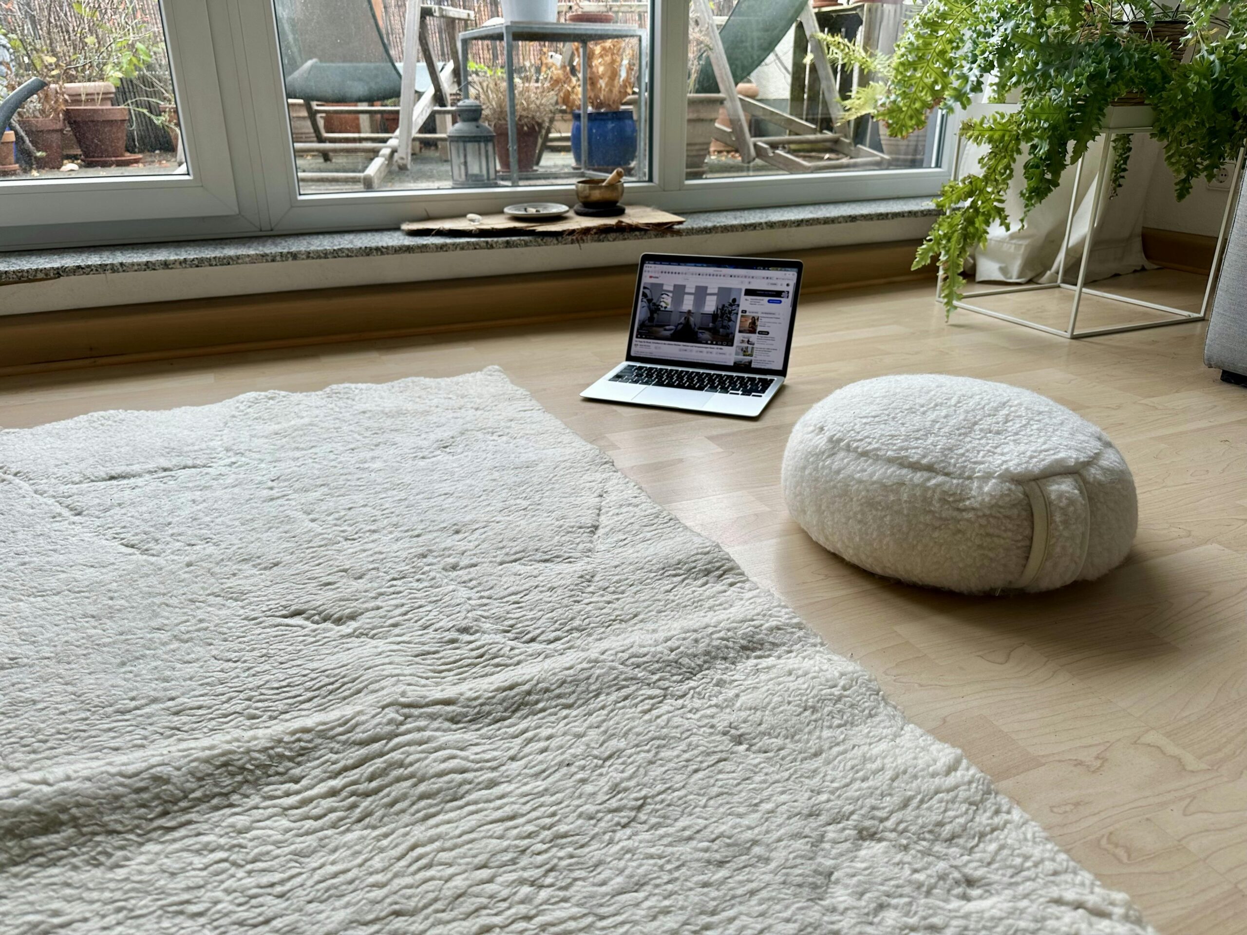 Schurwoll-Yogamatte auf dem Boden. Daneben ein Schurwoll-Meditationskissen sowie ein Laptop, auf dem ein Yogavideo zu sehen ist.