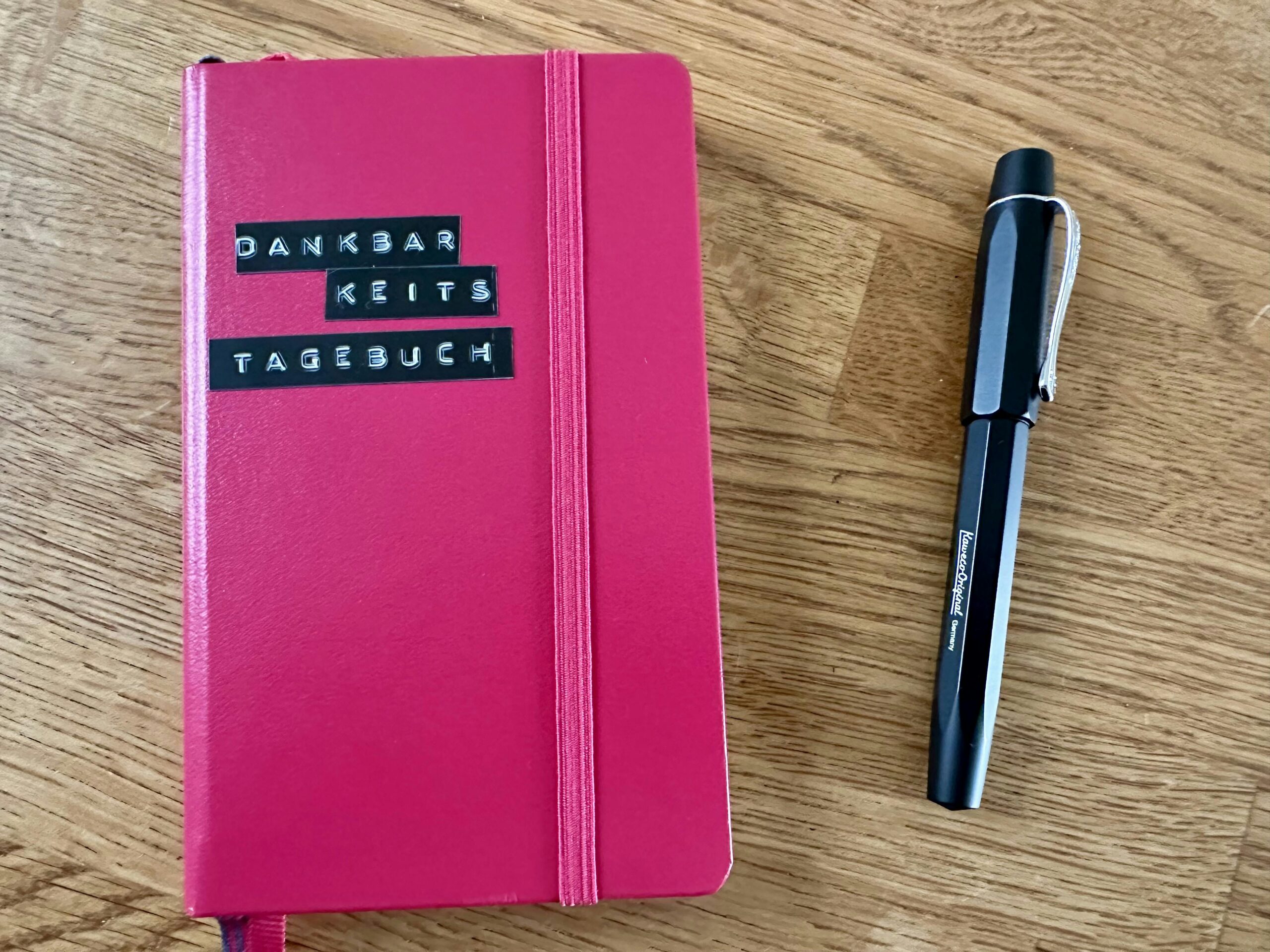 DIN A6 Notizbuch in pink, darauf ein schwarzes Klebeetikett "Dankbarkeitstagebuch". Daneben ein schwarzer Füller.