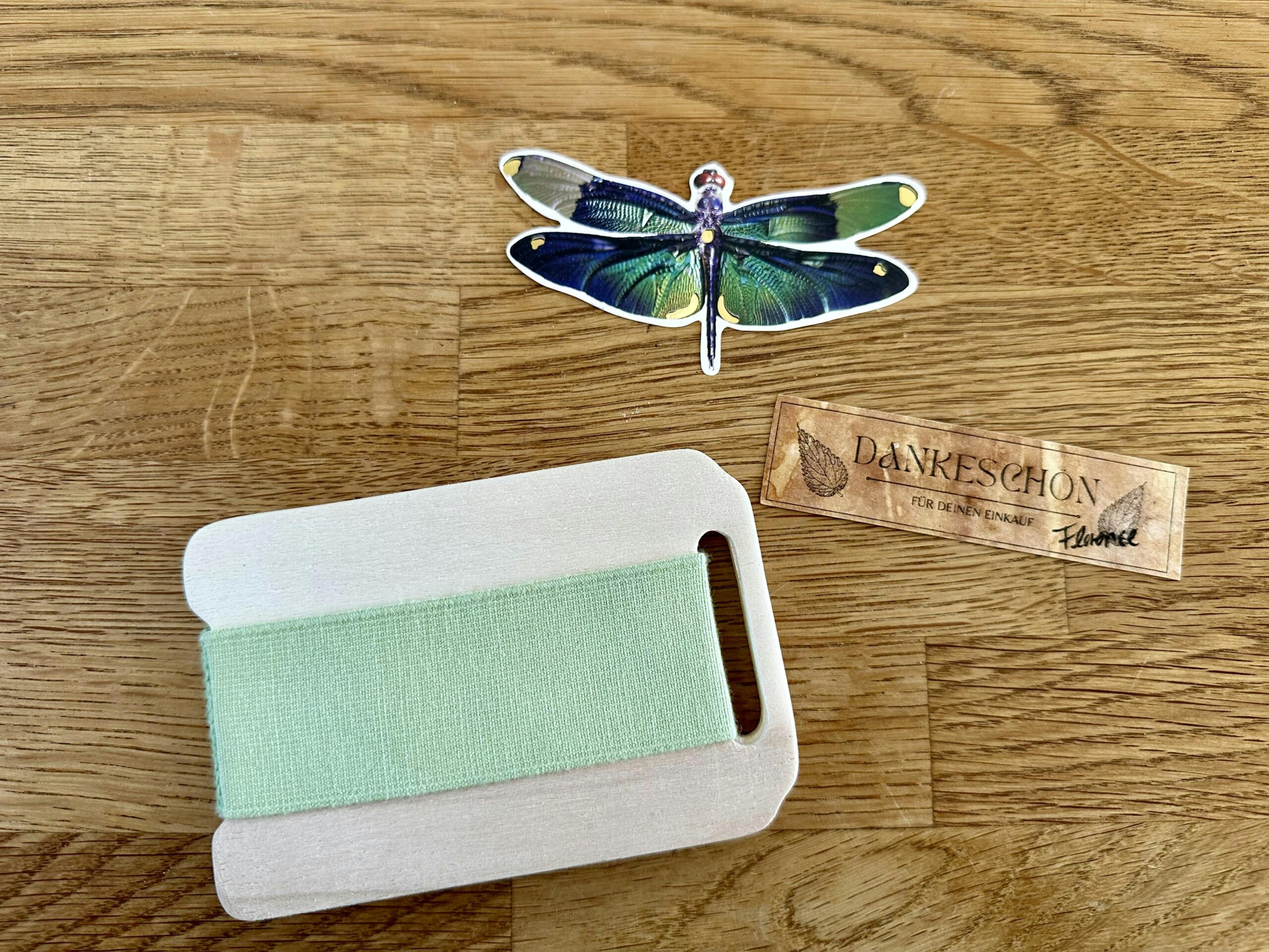 Kleine Pflanzenpresse aus Holz mit einem grünen Gummiband drumherum. Daneben ein Libellen-Sticker und ein brauner Zettel, auf dem Dankeschön" steht