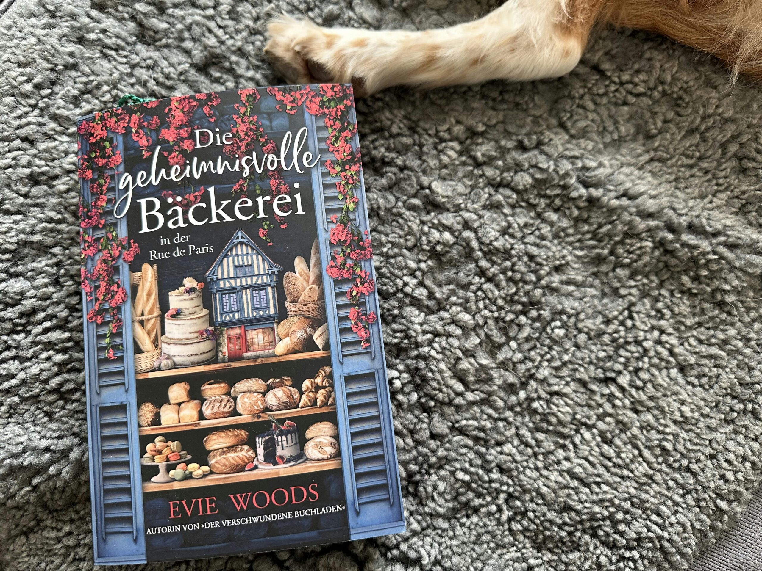 Buch "Die geheimnisvolle Bäckerei in der Rue de Paris" auf einem grauen Fell liegend. Daneben ragt das Bein eines Hundes ins Bild.