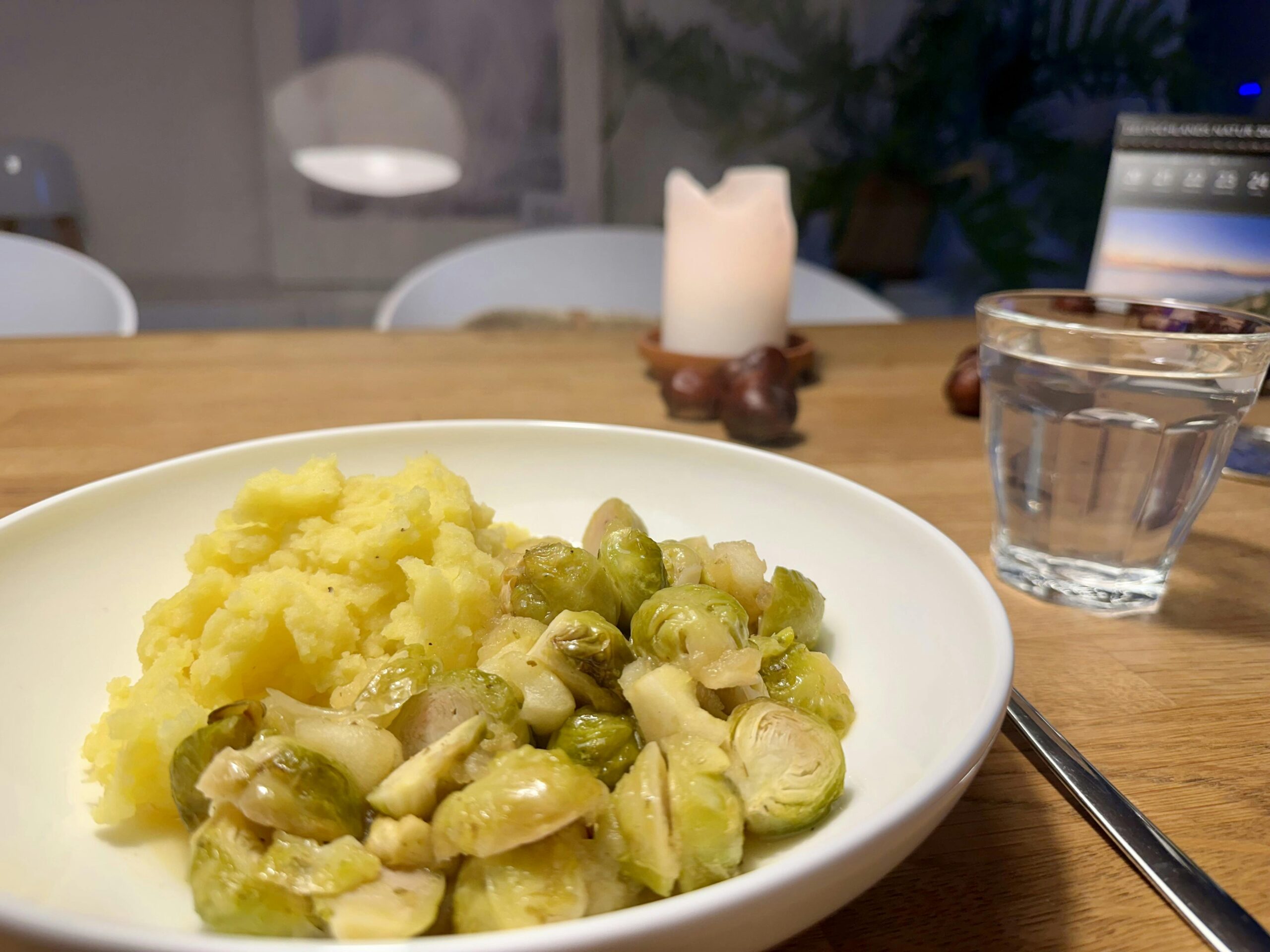Weißer, tiefer Teller mit Kartoffelpürree und Rosenkohl. Im Hintergrund brennt eine weiße Stumpenkerze und neben dem Teller steht ein Glas Wasser.