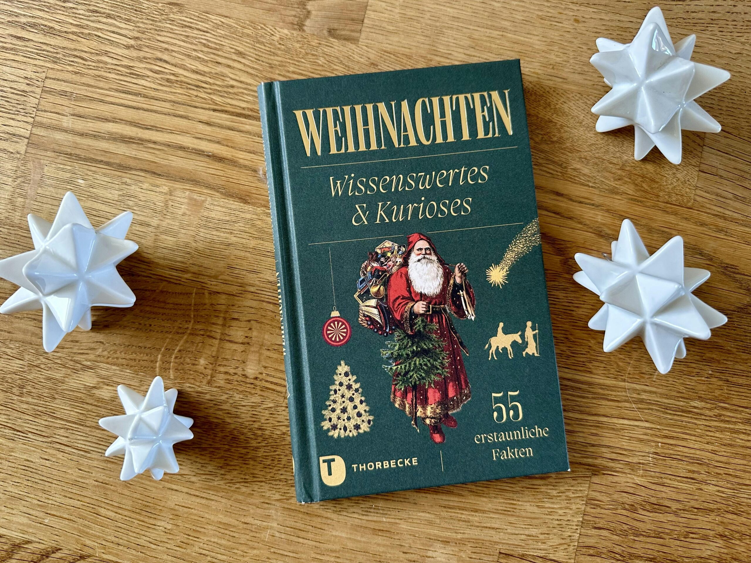 Buch "Weihnachten - Wissenswertes & Kurioses" auf einem Holztisch. Daneben vier weiße Porzellan-Sterne.