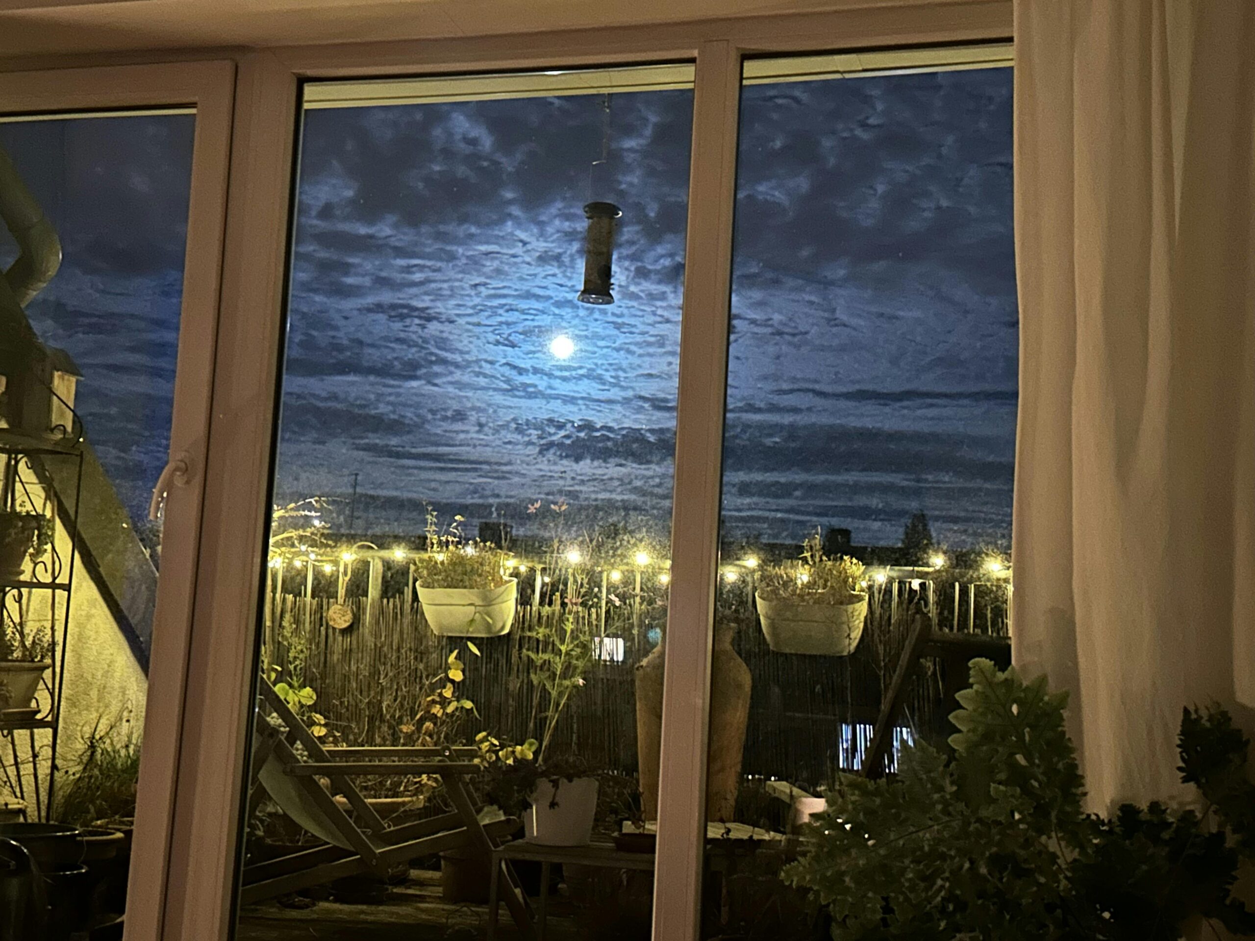Blick auf eine Terrasse mit brennender Lichterkette am Geländer und dem fast noch vollen Mond am Himmel.