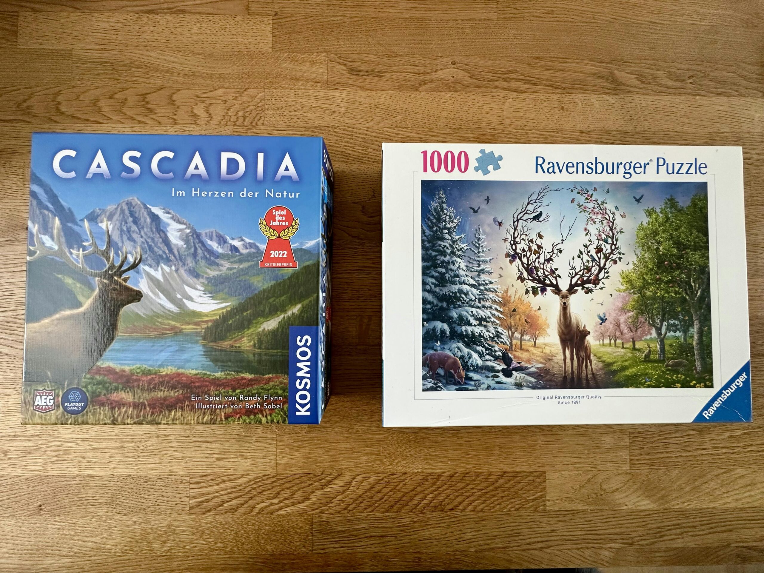 Der Karton vom Brettspiel "Cascadia" und von einem 1000-Teile Puzzle auf einem Holztisch. Auf beiden Kartons sind Hirsche zu sehen.