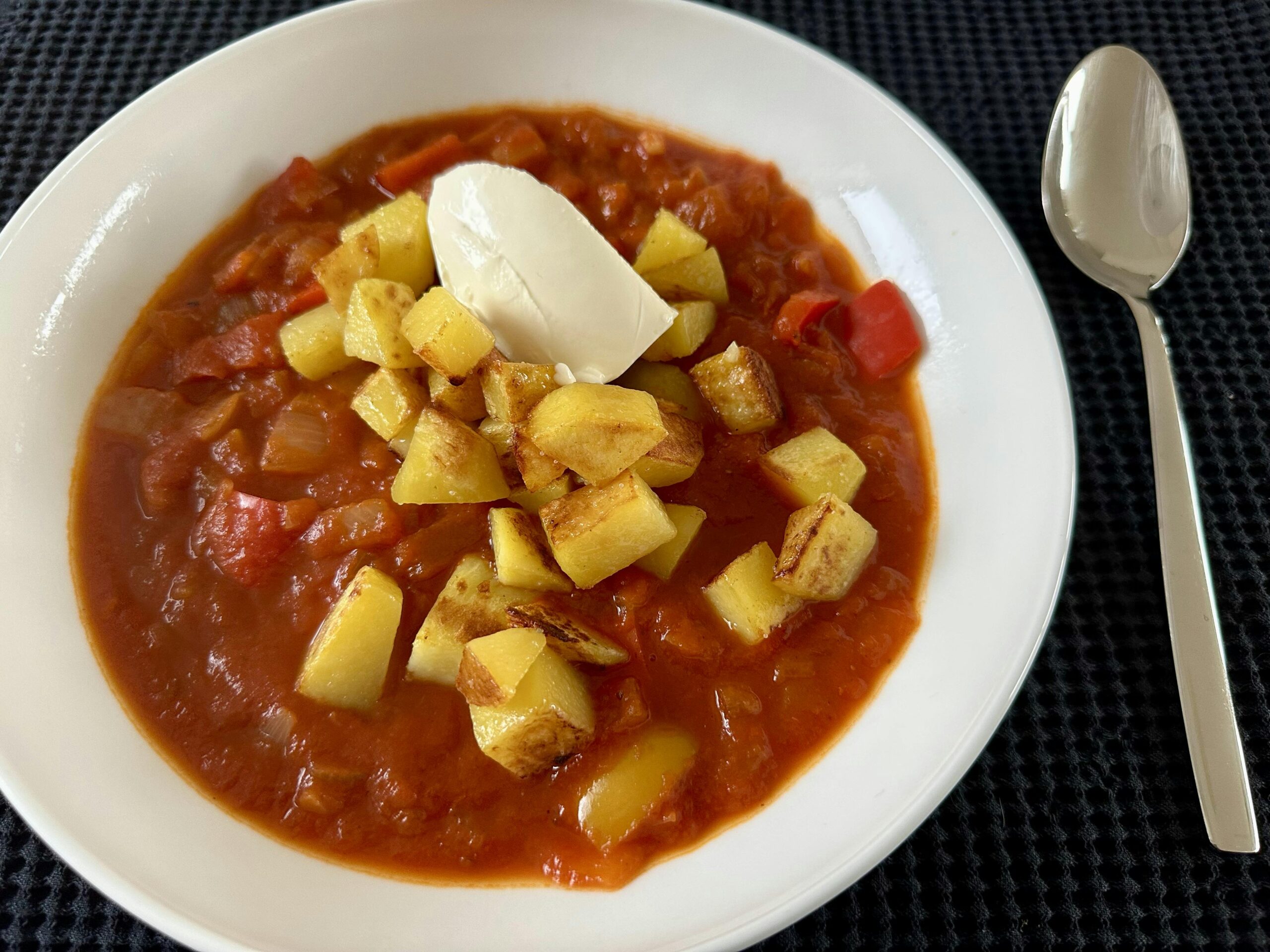Ein weißer tiefer Teller mit vegetarischem Gulasch mit Bratkartoffelwürfeln und einem Klecks Crème fraîche obendrauf.
