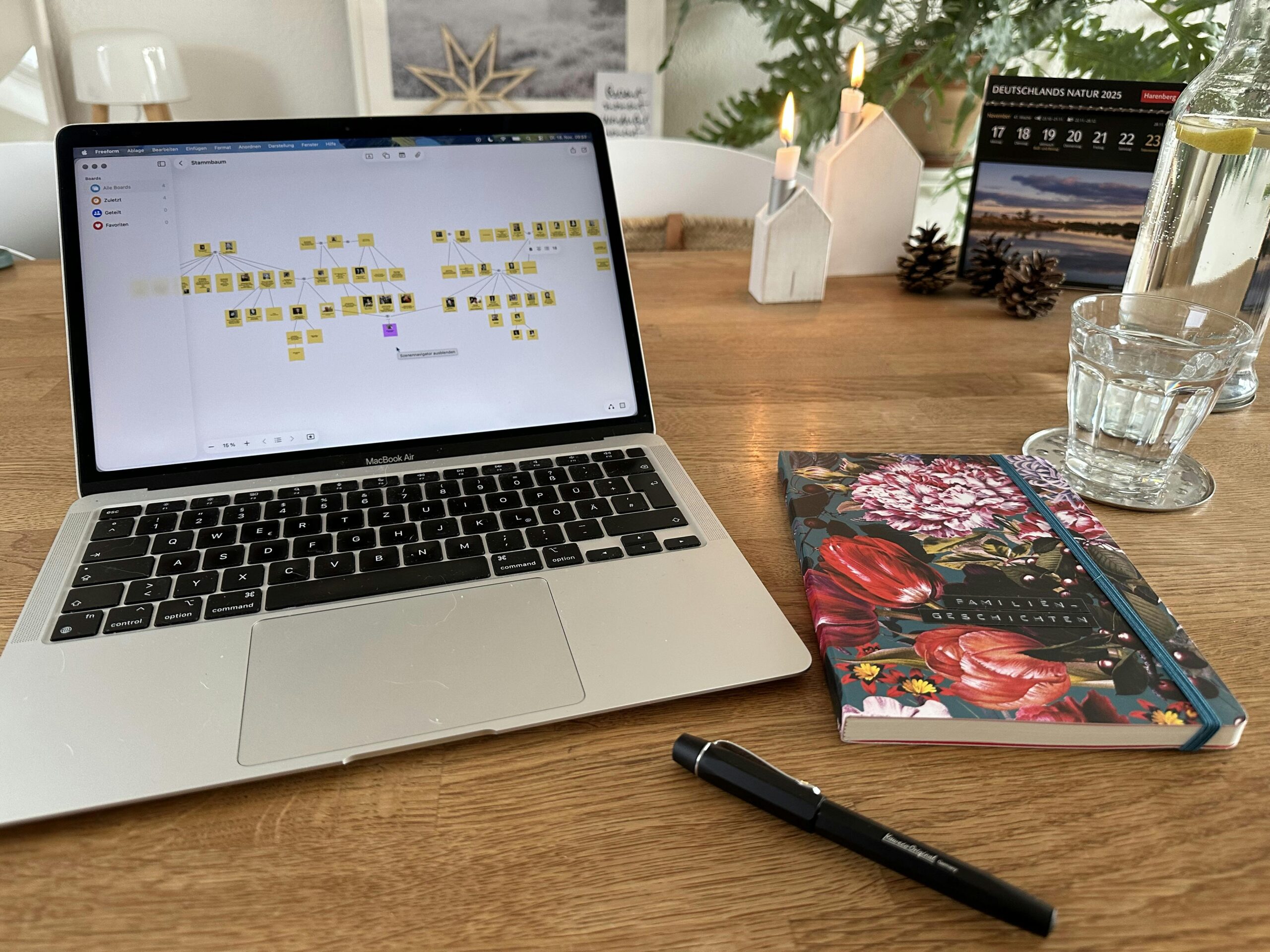 Auf einem Holztisch steht ein Laptop. Darauf sieht man eine Darstellung eines Stammbaumes. Neben dem Laptop liegt ein Notizbuch mit bunten Blumen, auf dem "Familiengeschichten" steht. Daneben ein schwarzer Füller.