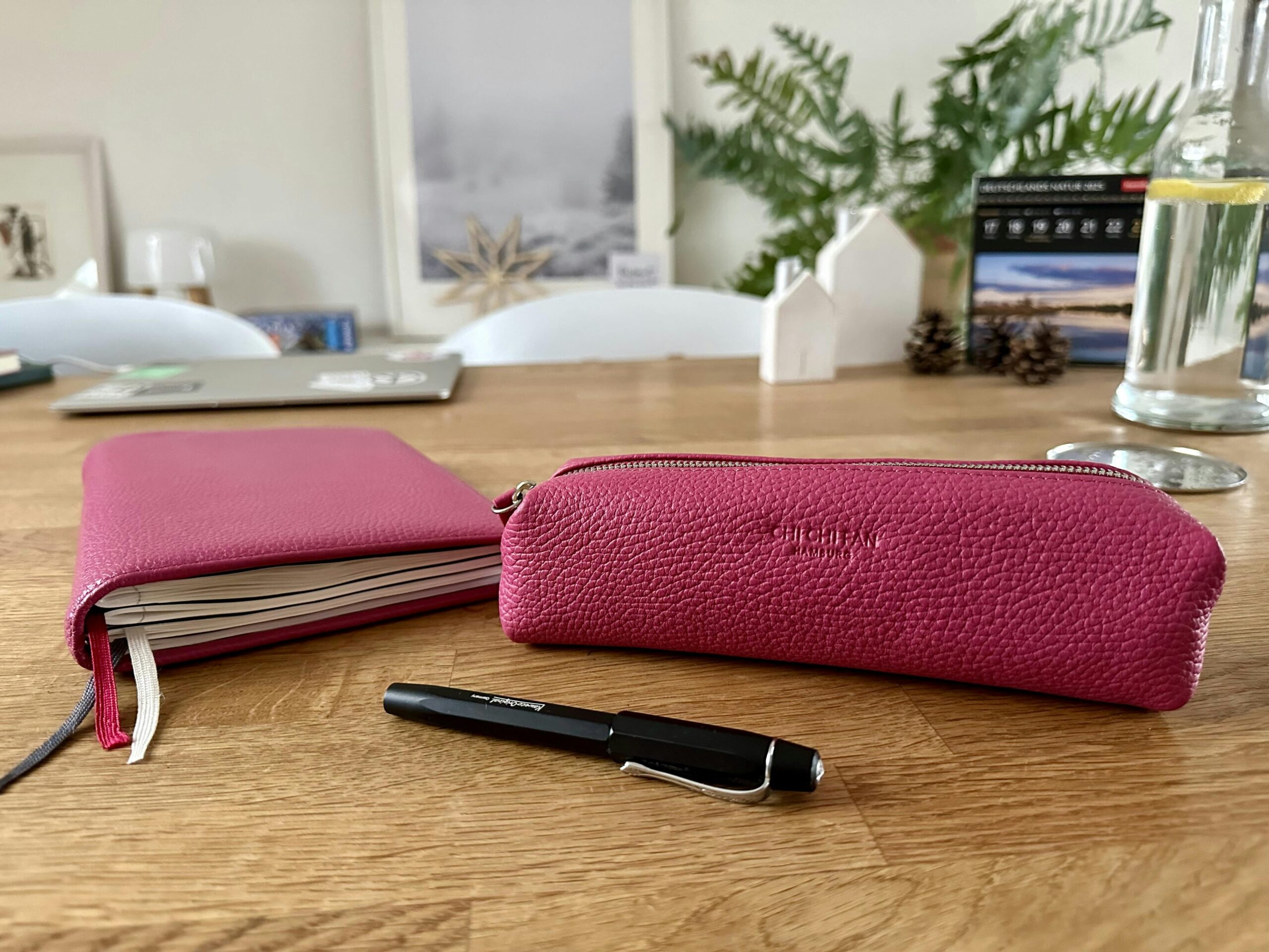 Auf einem Holztisch: Eine Lederhülle aus pinkfarbenem Leder mit Kalendereinlagen sowie ein passendes Leder-Stifteetui daneben. Dabei ein schwarzer Füller.