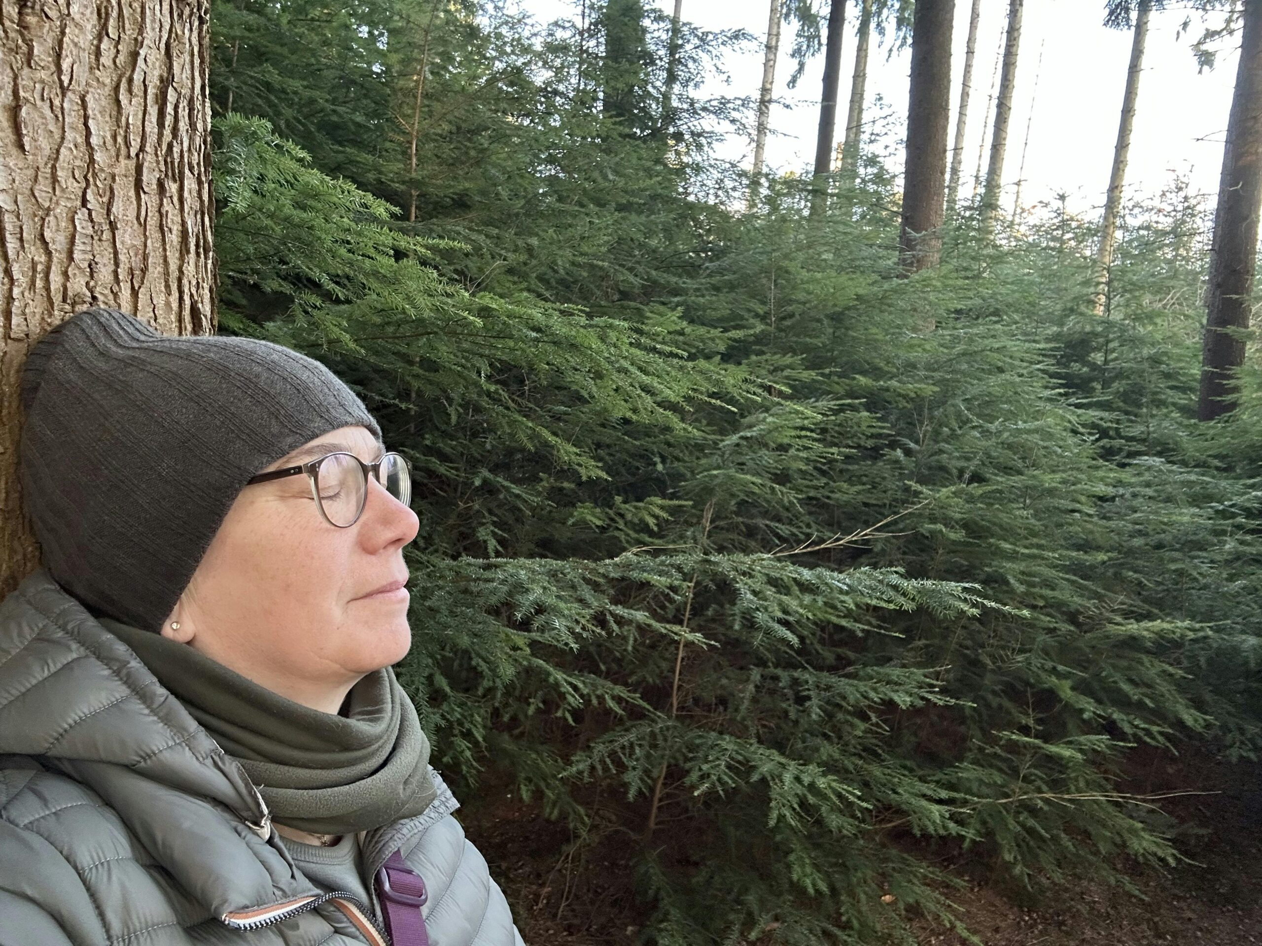Frau mit Mütze lehnt mit geschlossenen Augen an einem Baum im Wald.
