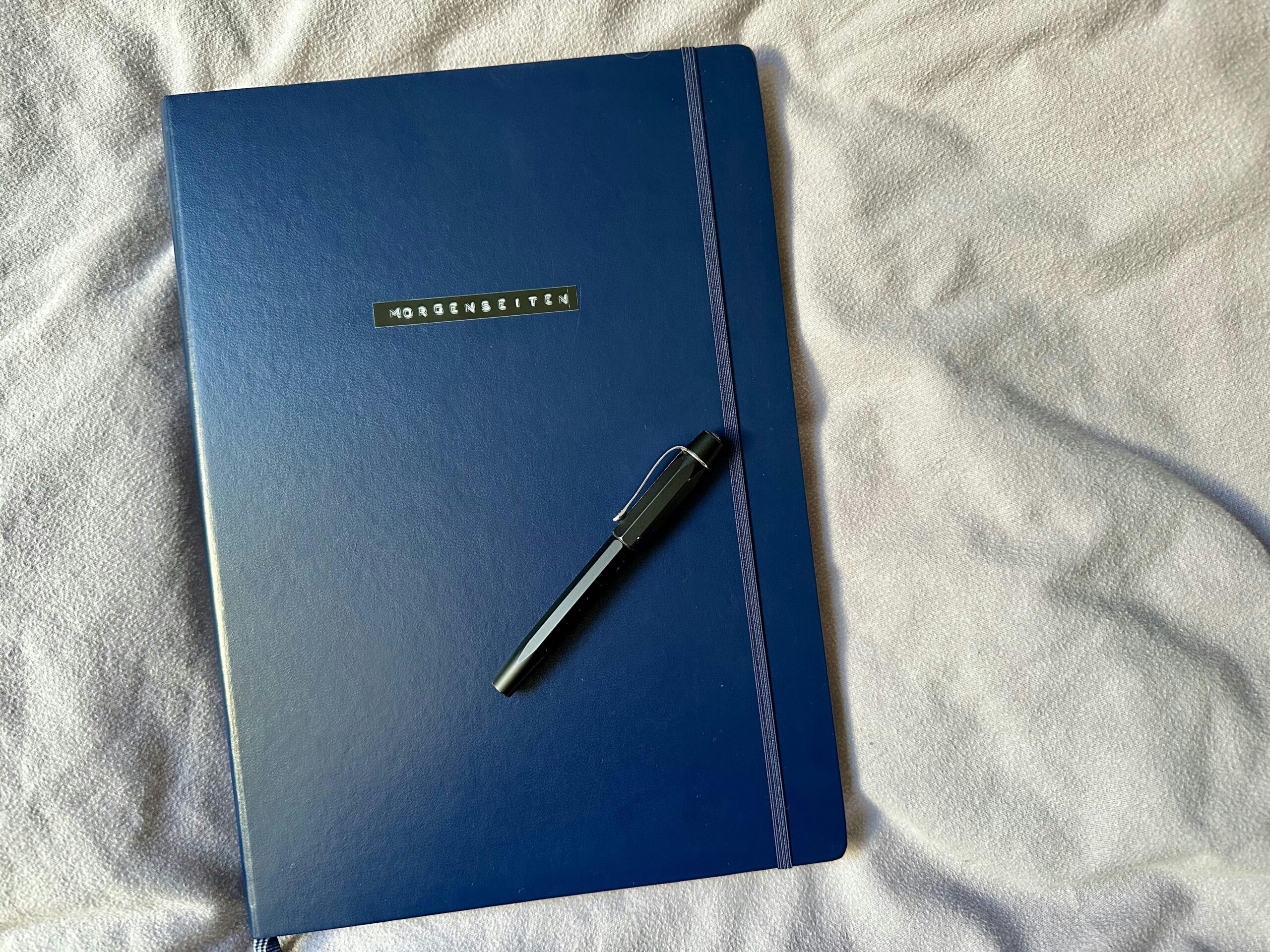 Dunkelblaue DIN A4 Kladde mit dem Aufdruck "Morgenseiten". Darauf ein schwarzer Füllfederhalter.