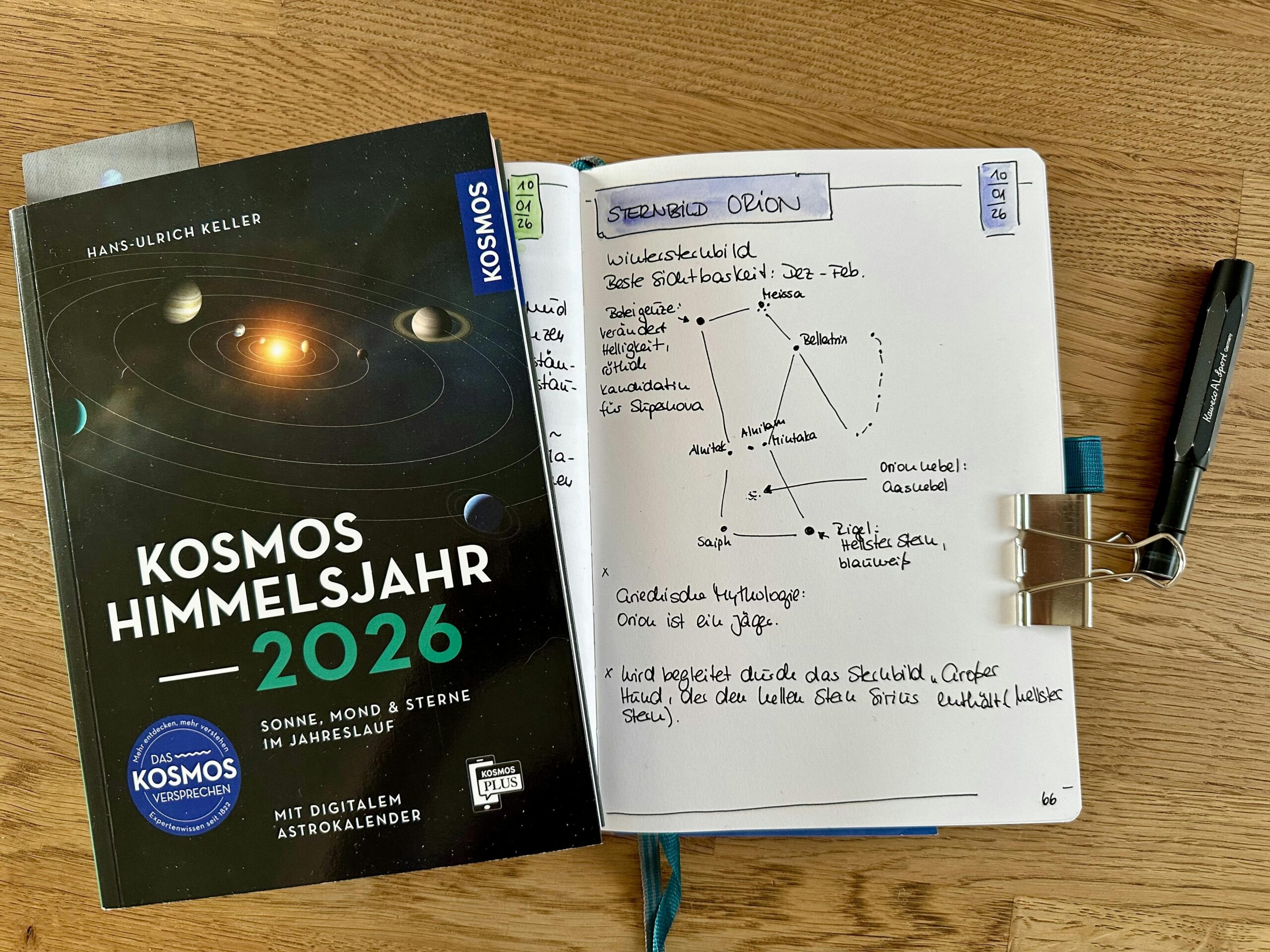 Buch "Kosmos Himmelsjahr 2026 und darunter schaut ein Notizbuch hervor mit einer Seite zum Sternbild Orion. Darauf finden sich Informationen sowie eine Skizze zu den Sternen.
